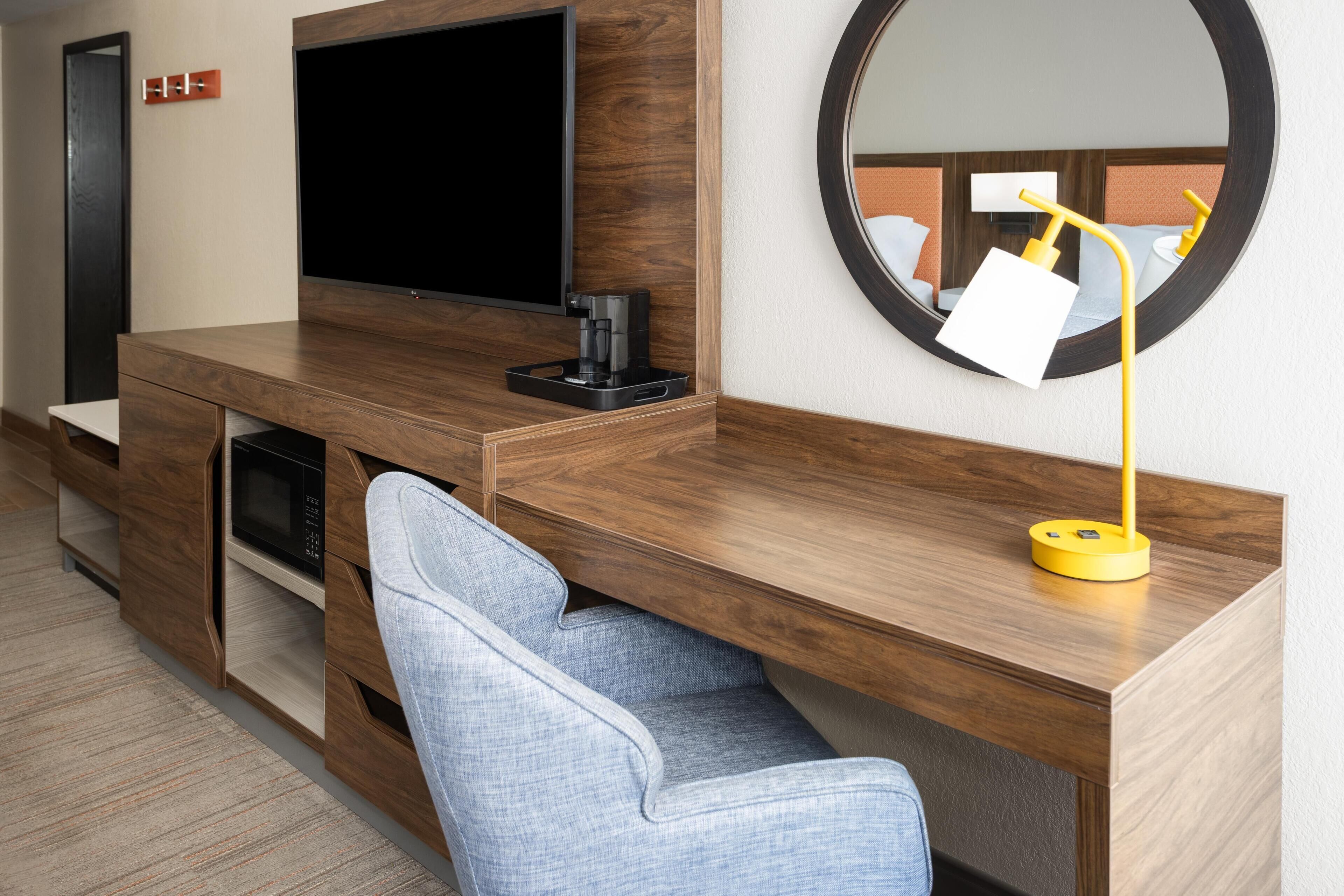 A modern hotel room with a wooden desk, a flat-screen TV, a coffee maker, and a round mirror.