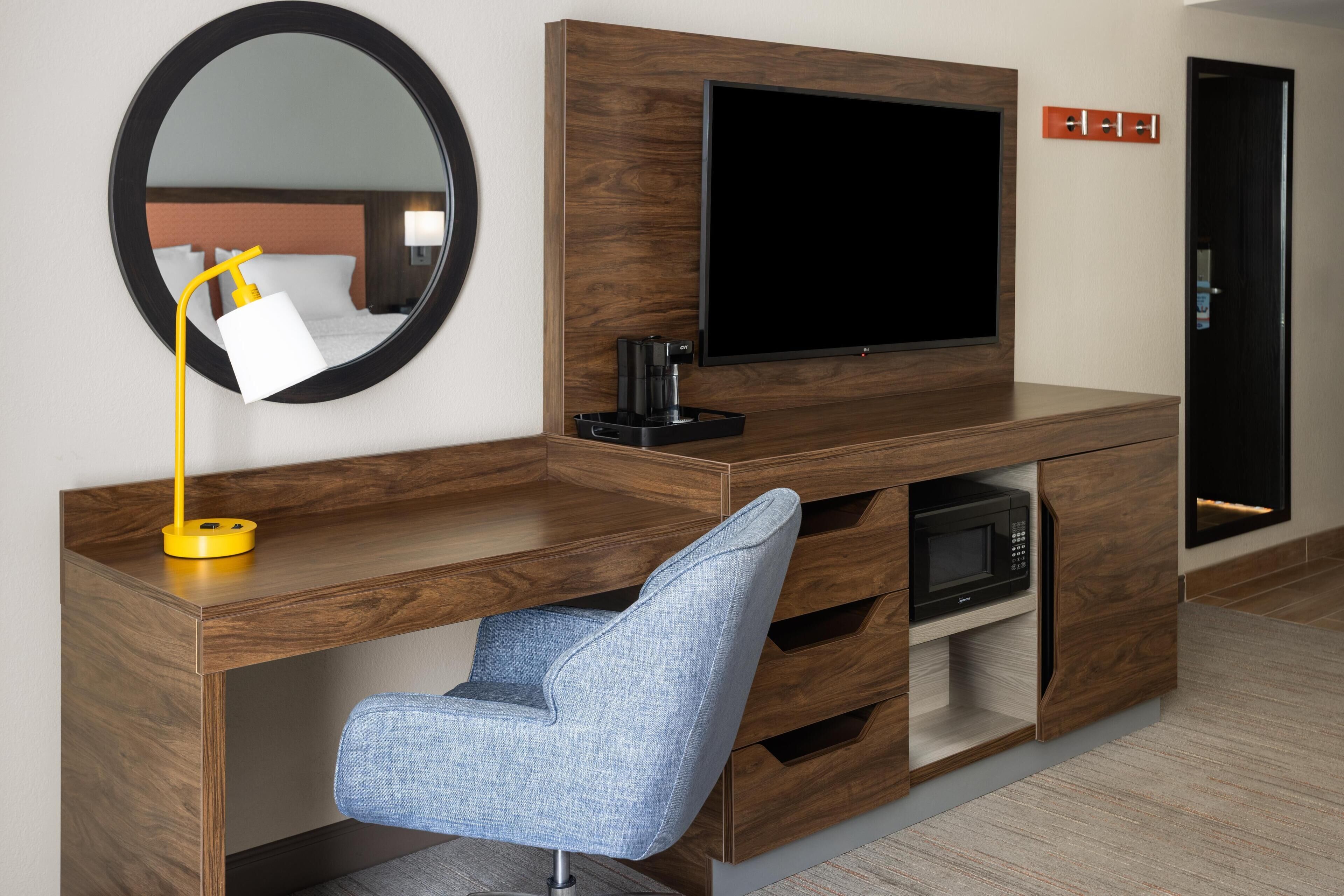 A modern hotel room with a wooden console desk, a blue chair, a flat-screen TV, a coffee maker, and a microwave.