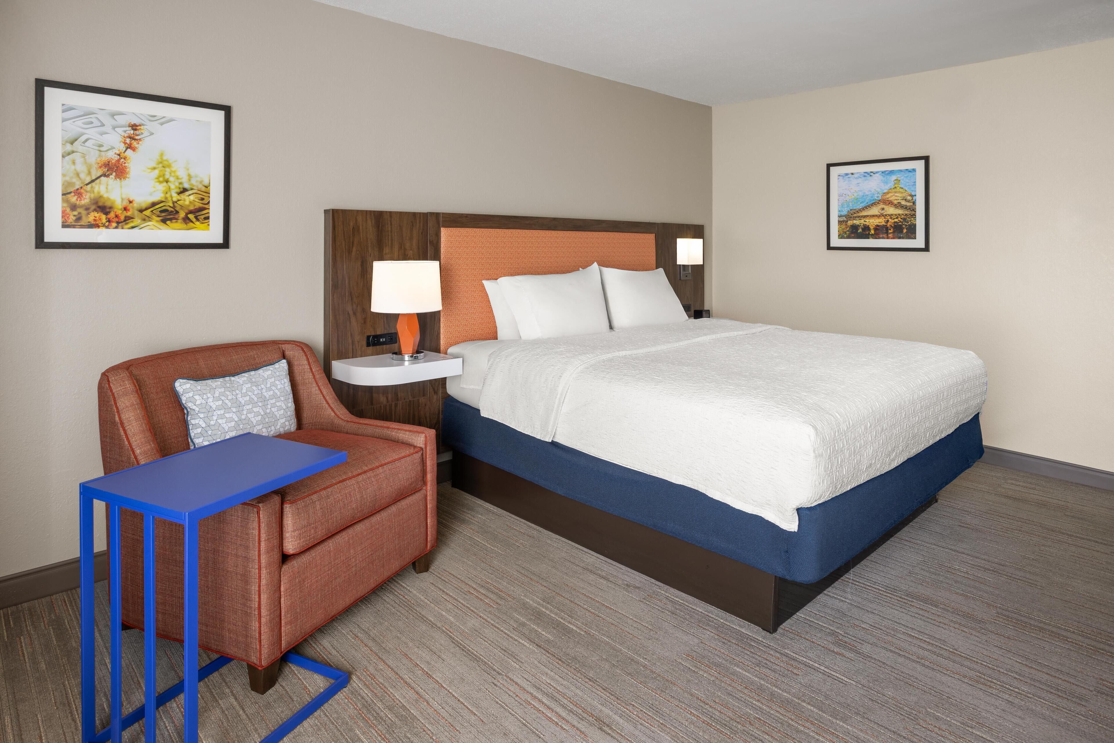 A hotel room with a bed, a nightstand, a lamp, a chair, and two framed pictures on the walls.