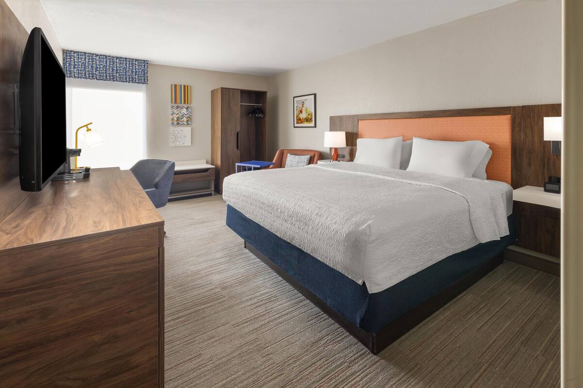 A hotel room with a large bed, a desk with a flat-screen TV, a chair, a wardrobe, and a nightstand with a lamp.