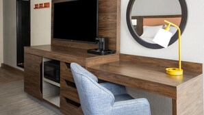 A modern hotel room with a wooden desk, a flat-screen TV, a microwave, a coffee maker, a round mirror, and a blue chair.