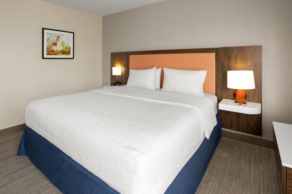 A hotel room with a large bed, a headboard, two bedside tables with lamps, a framed picture on the wall, and a small table by the bed.