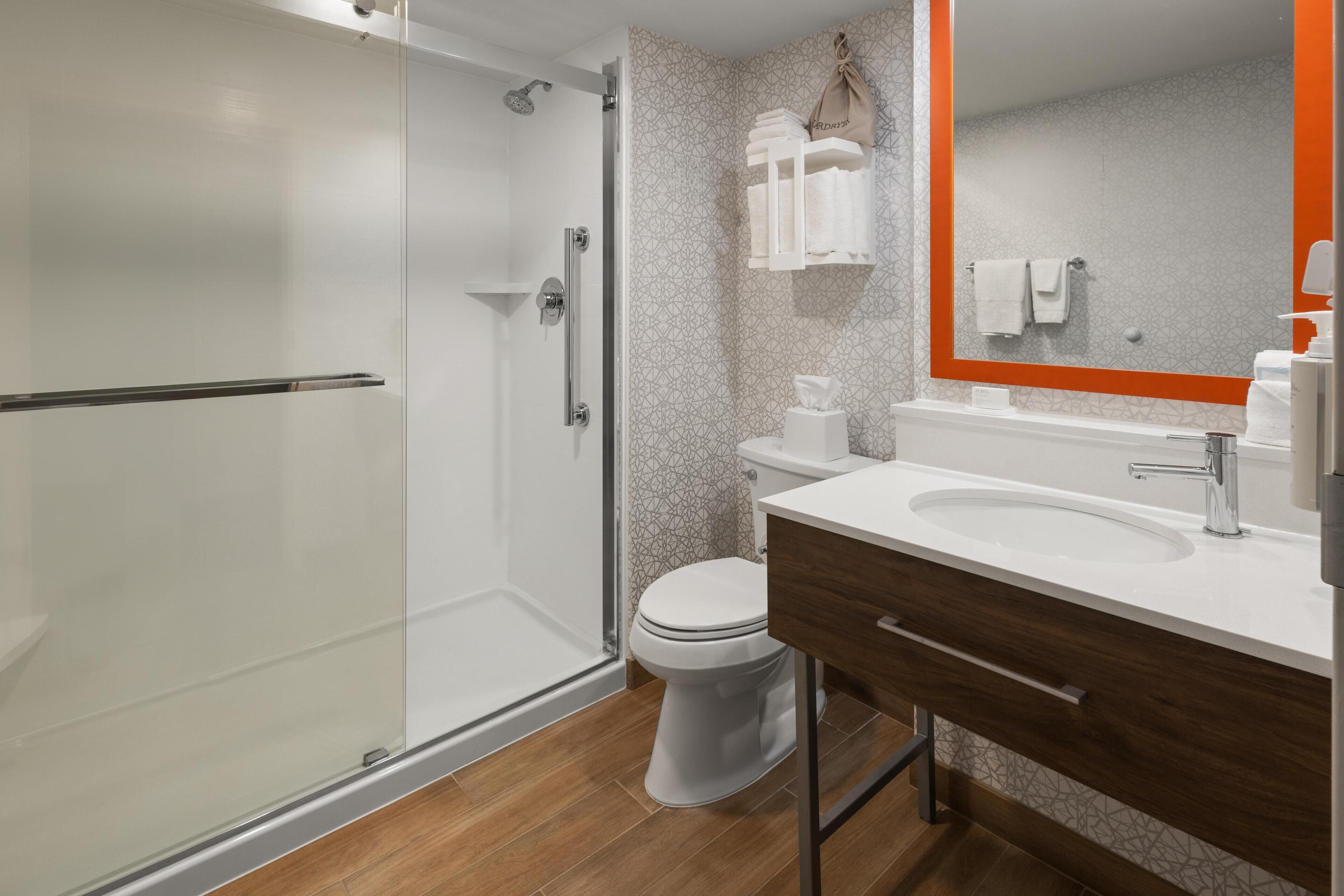 Un baño moderno con cabina de ducha de vidrio, inodoro blanco, tocador de madera con lavamanos y espejo con marco naranja.