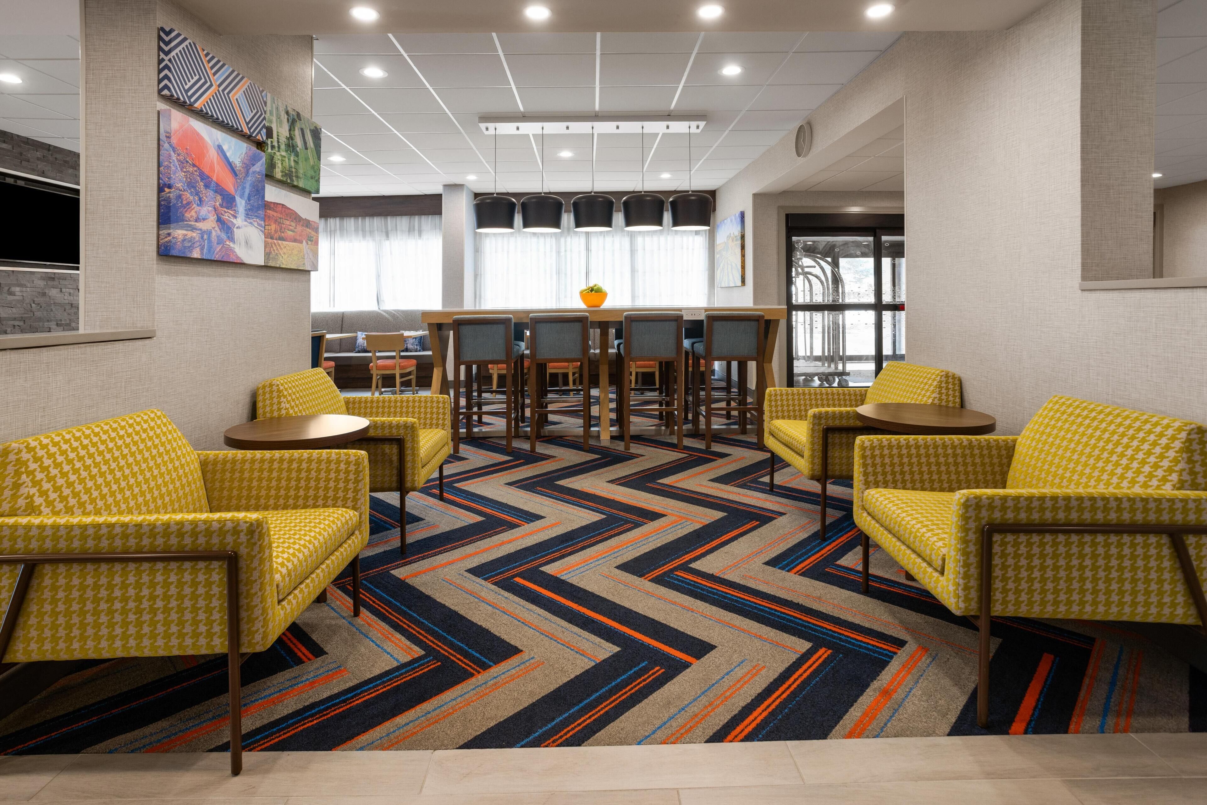 Un hall d’hôtel moderne avec un espace de restauration central, des chaises à motifs jaunes et une moquette multicolore.