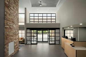 Zona de recepción con pared de piedra, puerta doble de entrada, mostrador de recepción y techo alto con iluminación empotrada.