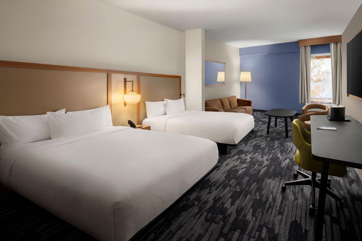 A hotel room with two beds, a desk, a chair, a lamp, and a window.