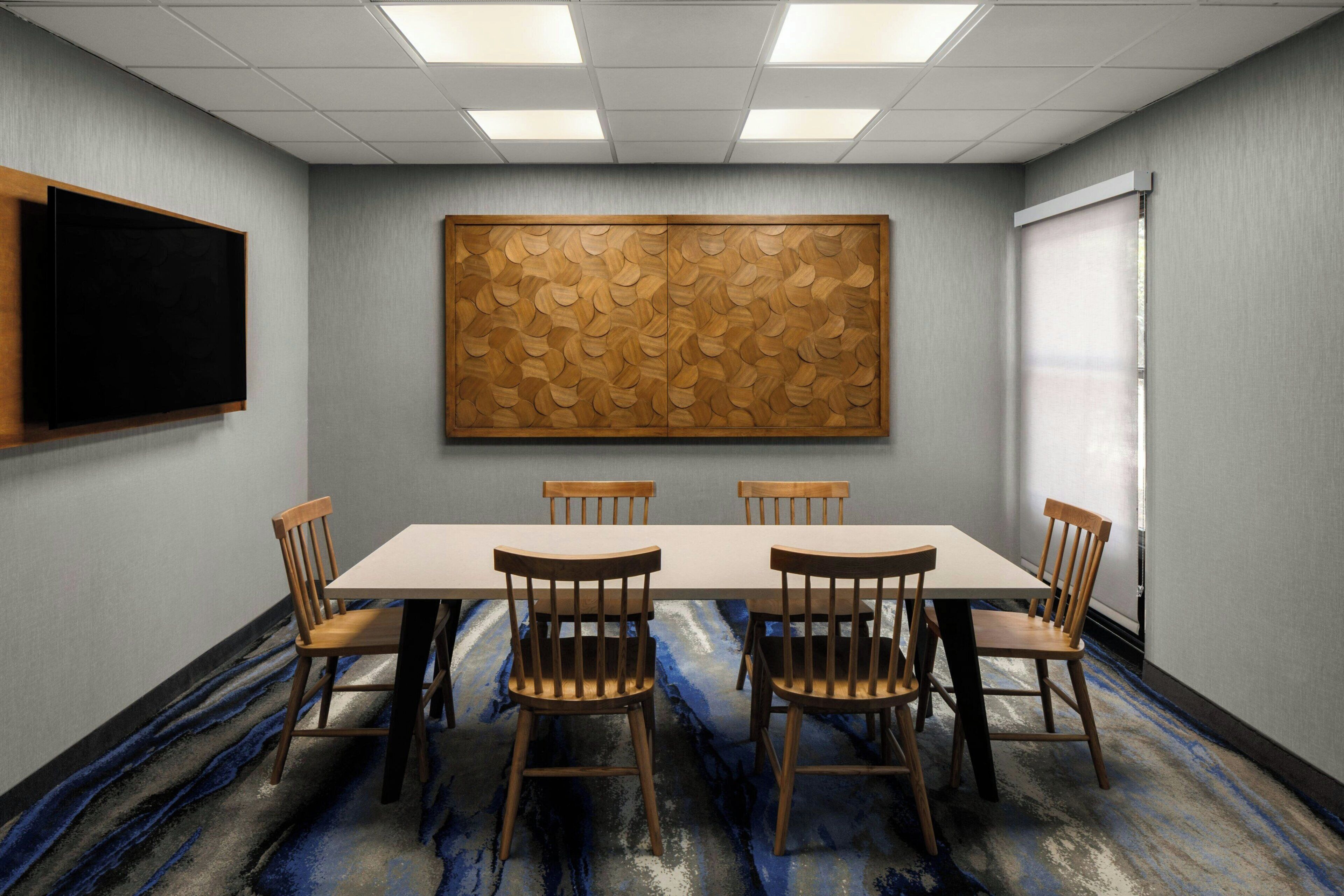 Een vergaderruimte met een lange tafel, zes stoelen, een groot houten paneel aan de muur, een schoolbord en een raam met jaloezieën.