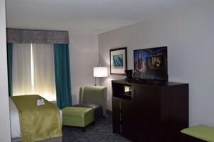 A hotel room with a bed, a green armchair, a television, a desk with a lamp, and a framed picture on the wall.