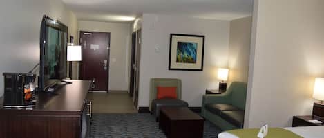Habitación de hotel con cama, escritorio con lámpara, silla, sofá, cafetera y un cuadro en la pared.