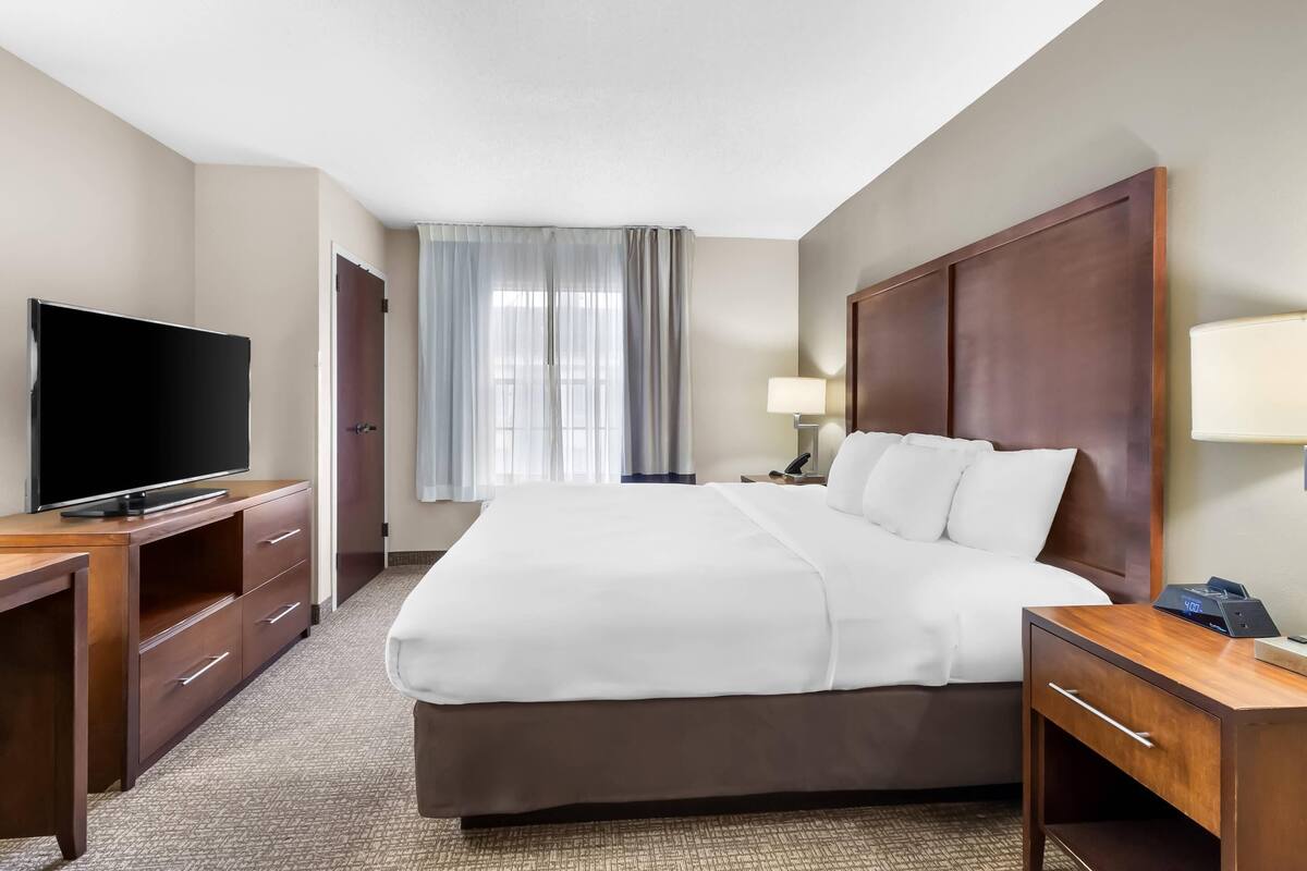 A hotel room with a large bed, a TV on a wooden desk, a nightstand with a lamp, and a window with curtains.