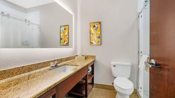 A bathroom with a granite countertop, a white toilet, a glass shower enclosure, and two framed artworks on the wall.