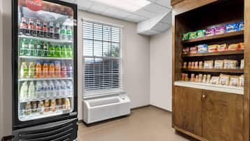 A vending machine with various drinks and snacks, a window with blinds, and a wooden cabinet with snacks.