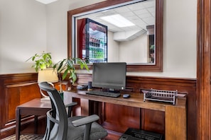 A hotel room with a wooden desk, a computer, a chair, a lamp, and a potted plant.