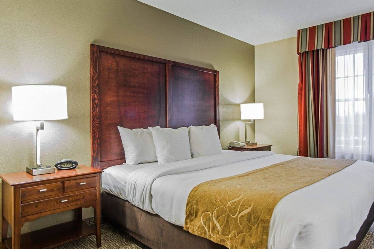 A hotel room with a wooden headboard, a bed with white linens, two bedside tables with lamps, and a window with curtains.