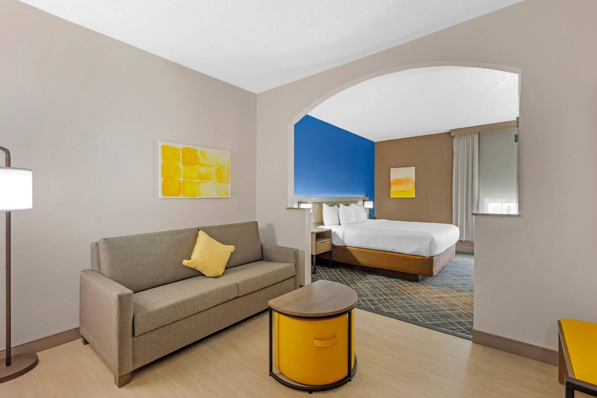 A hotel room with a sofa, a round yellow side table, a bed, and a large mirror.