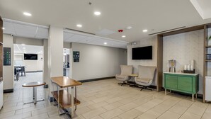 Un espace d’attente moderne avec une table en bois, deux chaises, un meuble vert et un téléviseur fixé au mur.