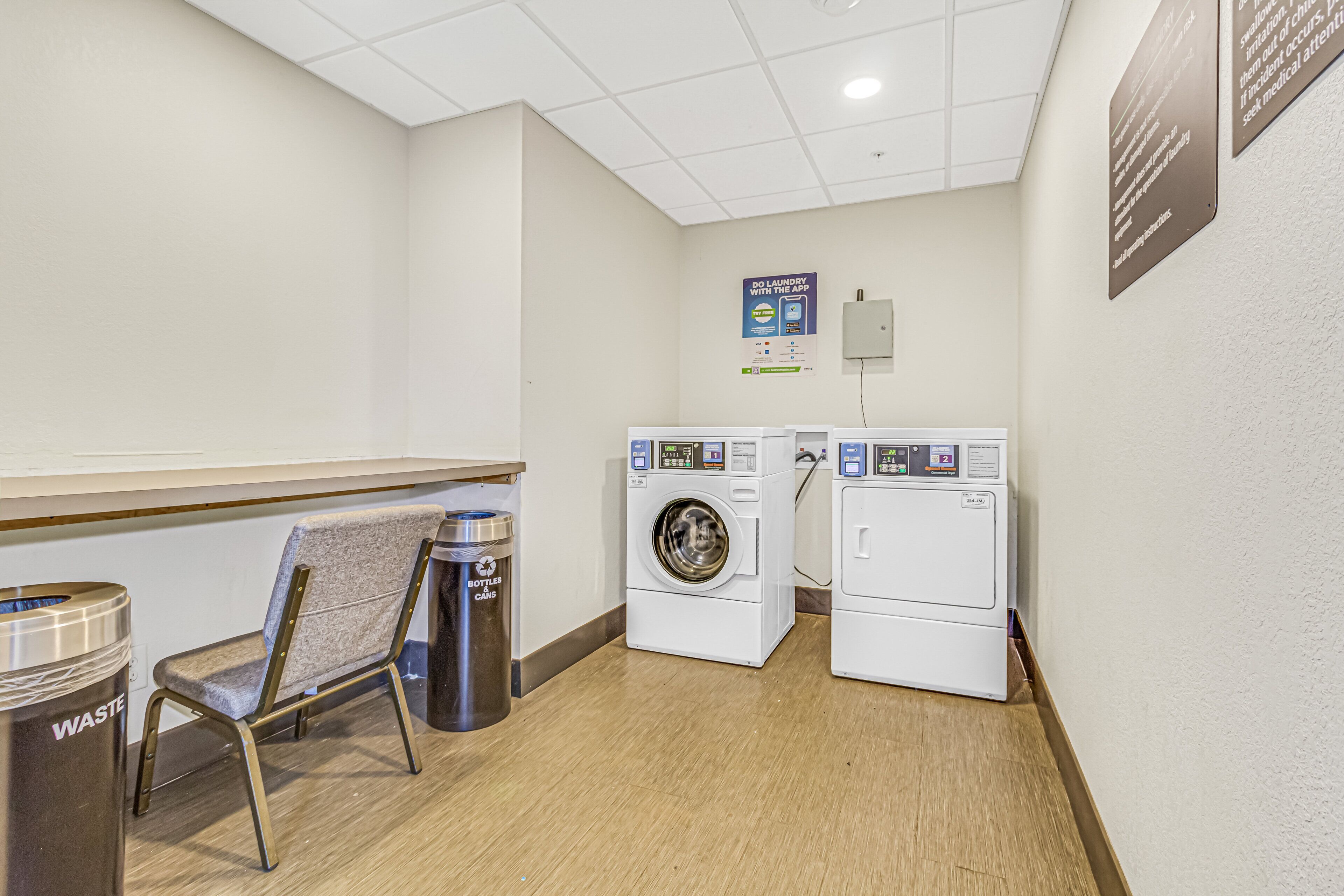 A laundry room with two washing machines, a chair, and a trash can.