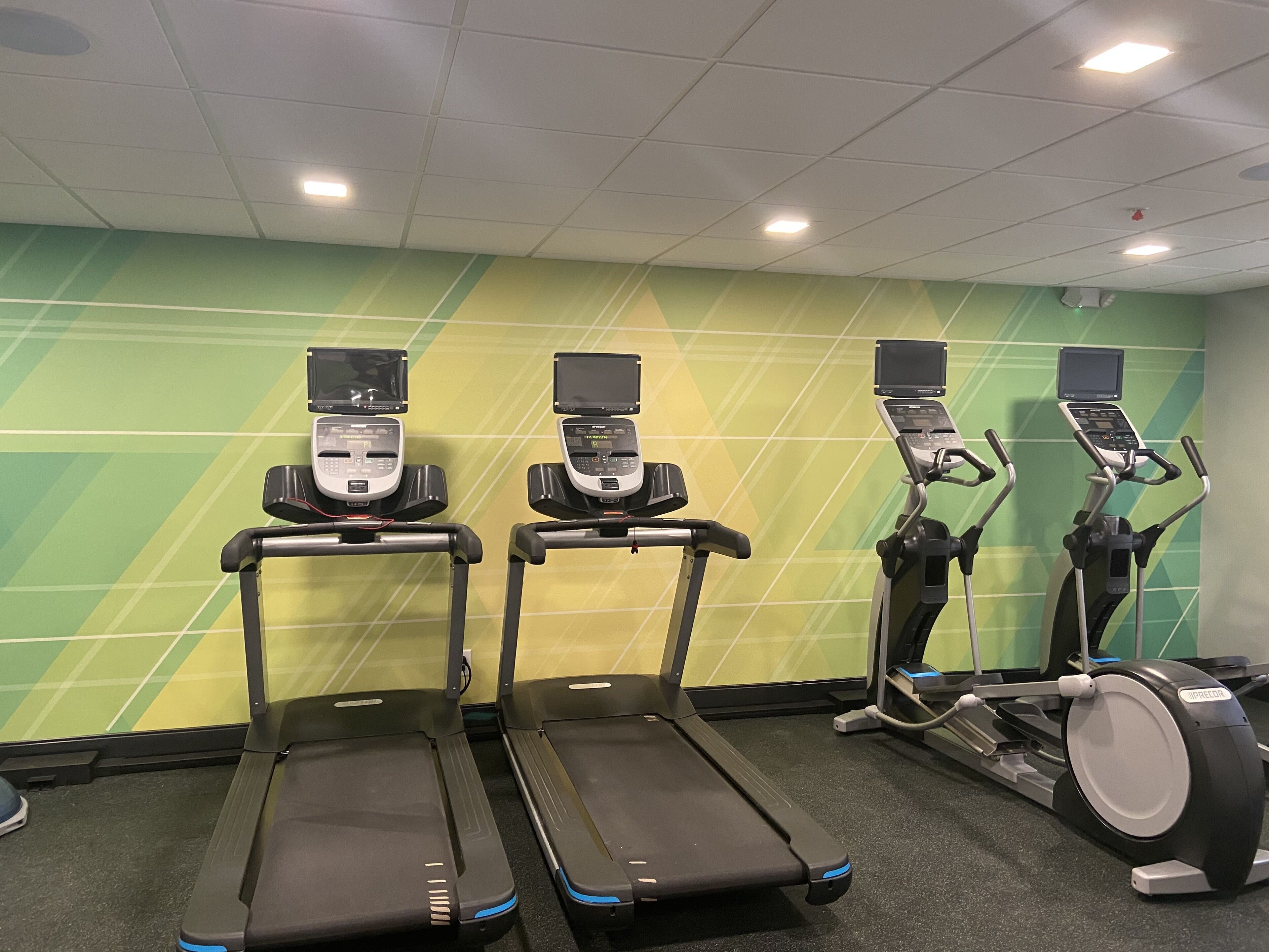 Drei Laufbänder und ein Crosstrainer in einem Fitnessraum mit grün-gelb gemusterten Wänden.