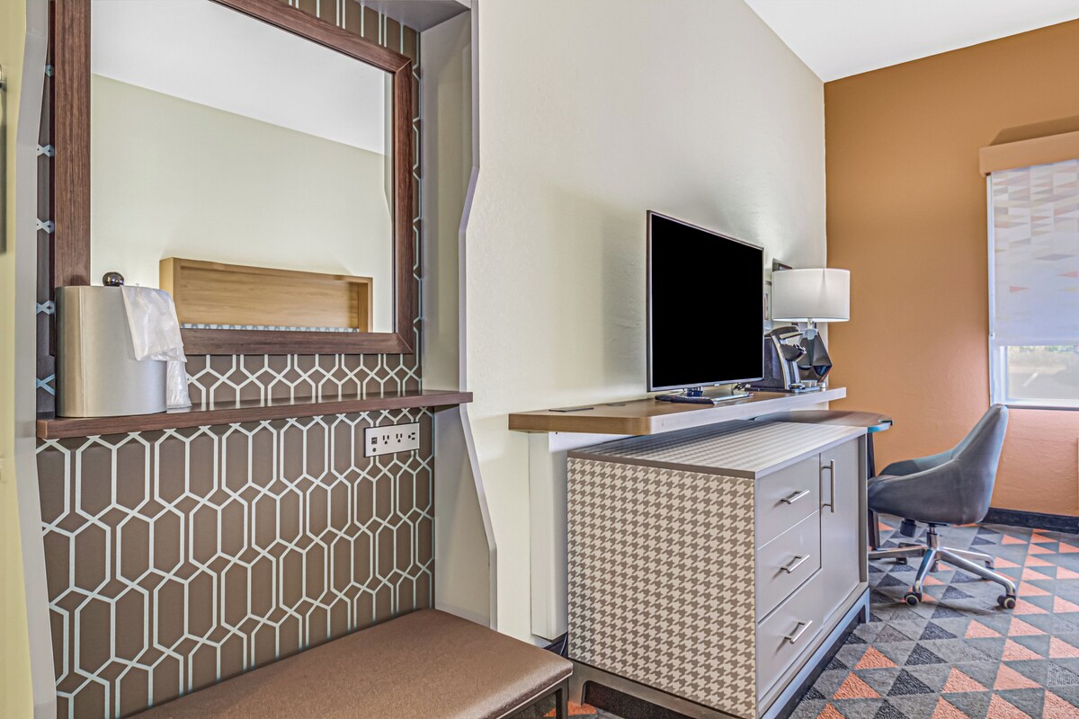 Ein Hotelzimmer mit einem Bett, einem Schreibtisch mit Computer, einem Sessel und einem Spiegel, sowie einer Wand mit Muster.