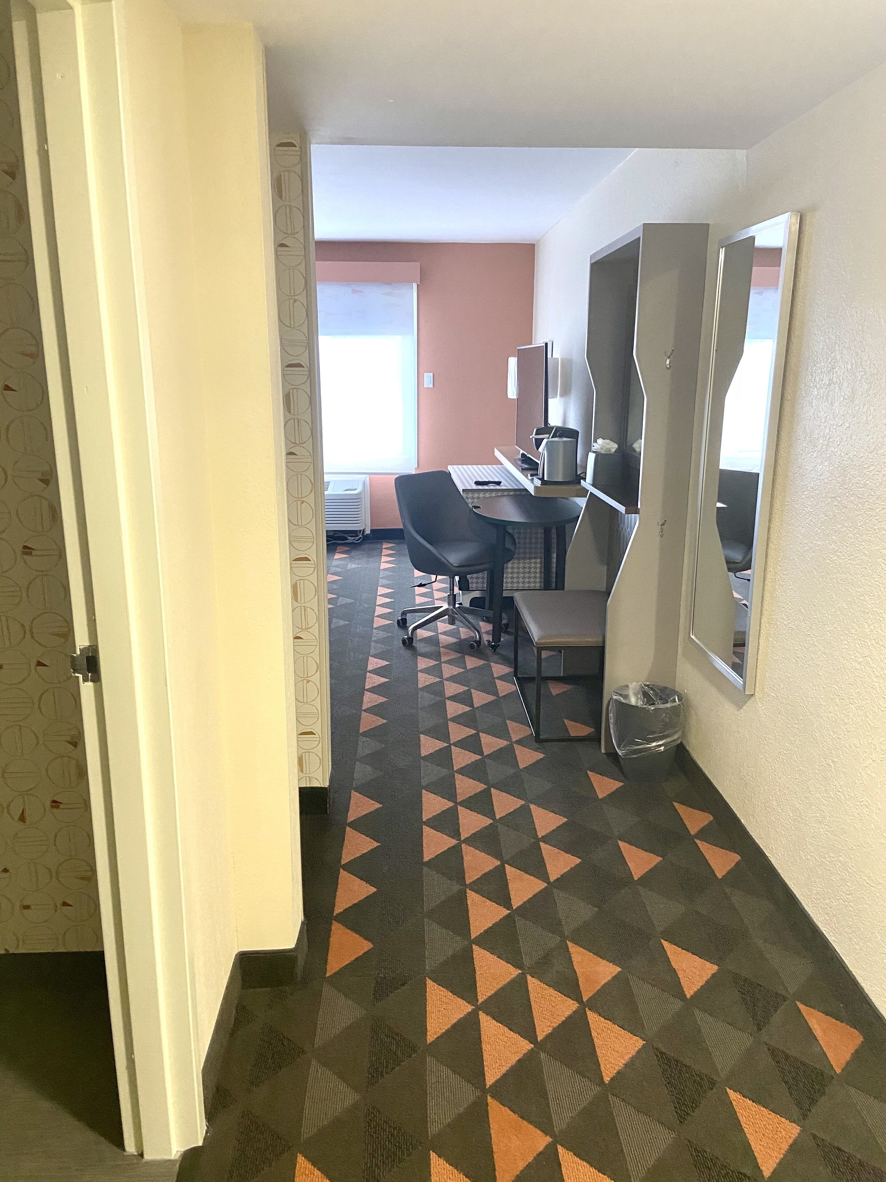 Habitación de hotel con escritorio, silla y ordenador. Se observa una ventana con persianas, un radiador y un cubo de basura.