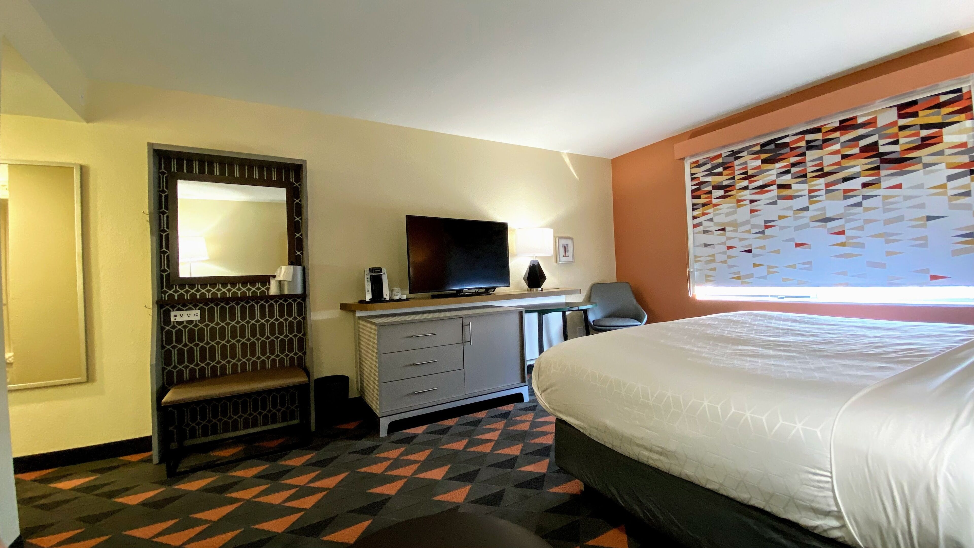 Habitación de hotel con cama, escritorio con televisor, silla y ventana con persianas decoradas.