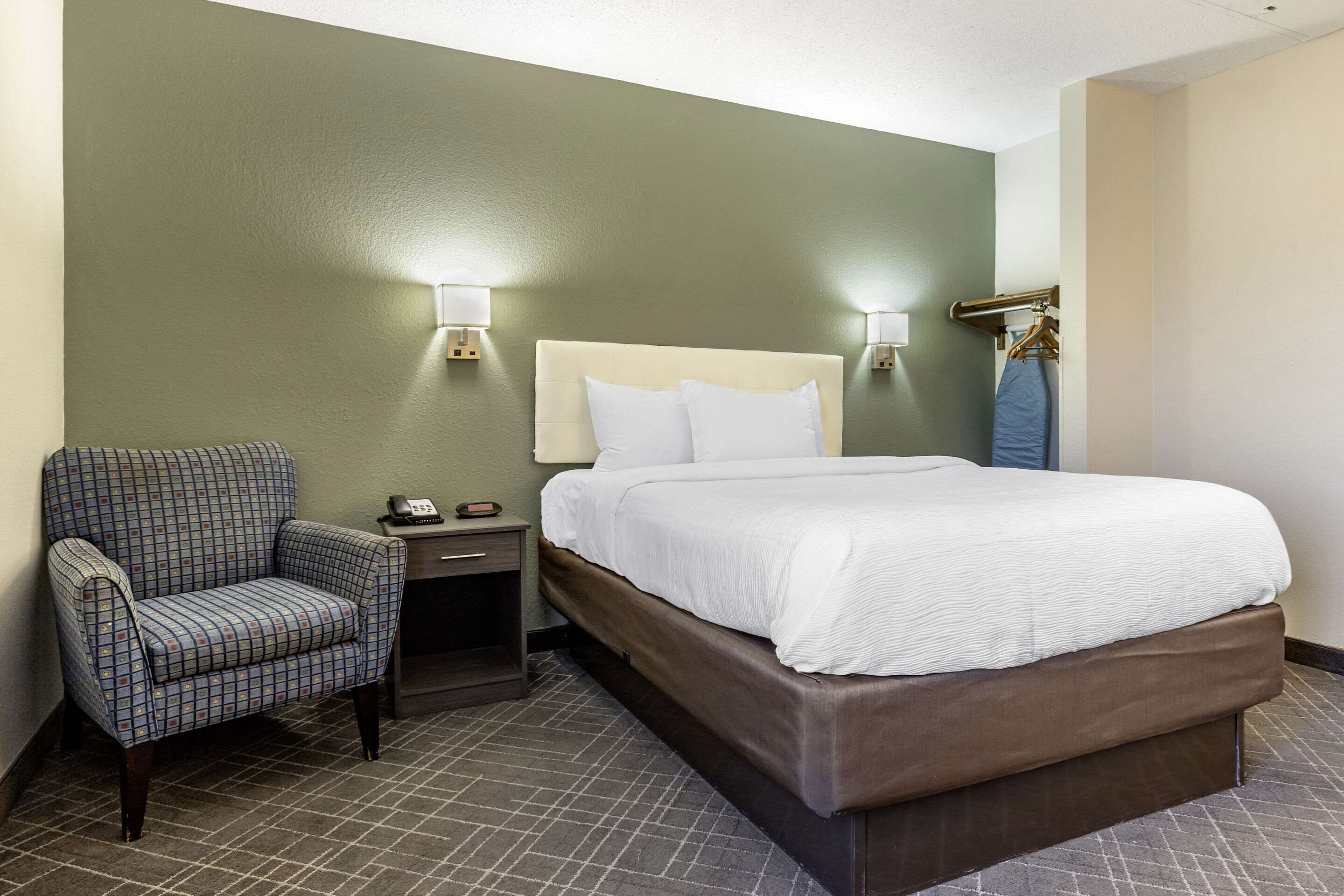 A hotel room with a bed, a chair, a nightstand, and a wall-mounted lamp.