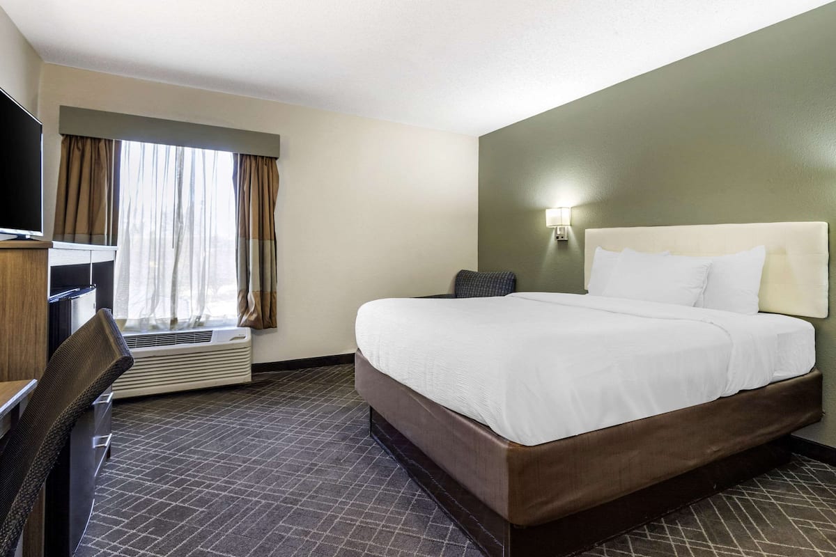 Ein Hotelzimmer mit einem großen Bett, einem Schreibtisch mit Stuhl, einem Fernseher und einem Fenster mit Vorhängen.
