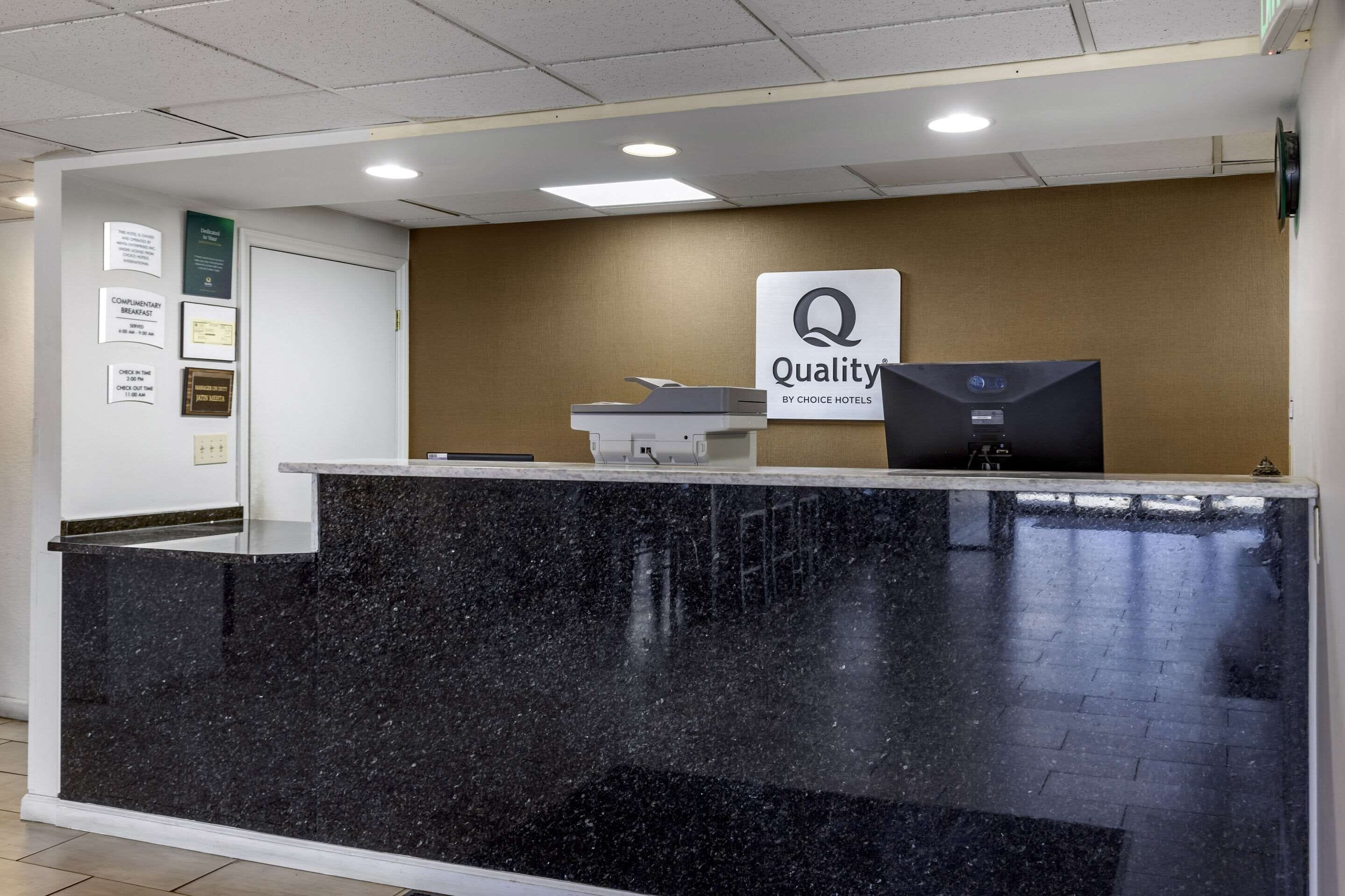 Un comptoir d’accueil d’hôtel avec un plan de travail en granit noir, un ordinateur et une enseigne indiquant « Qualité garantie par Choice Hotels ».