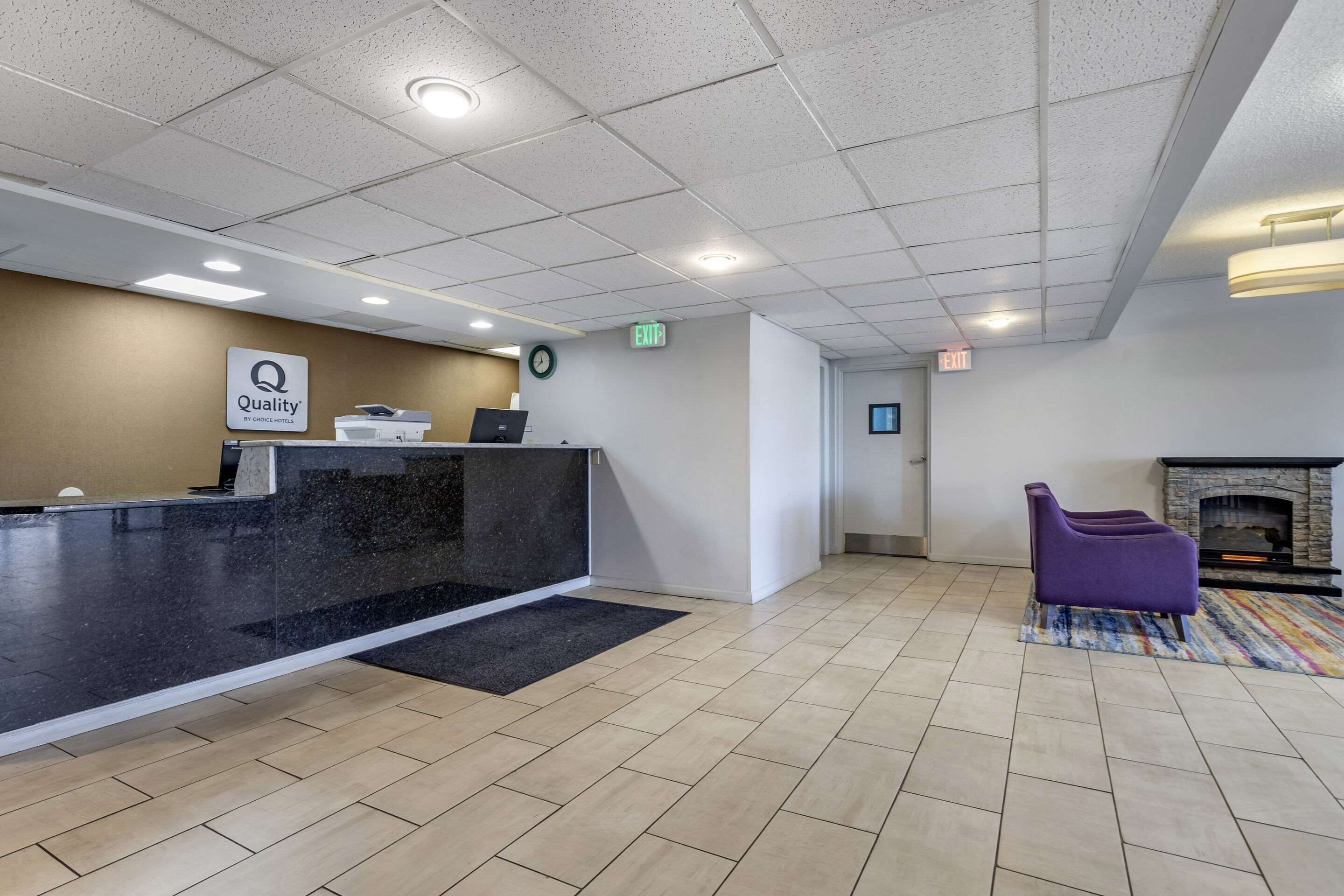 A reception area with a black reception desk, a sign reading "Quality by Caruso Hotels," a clock, and a fireplace with a purple armchair.