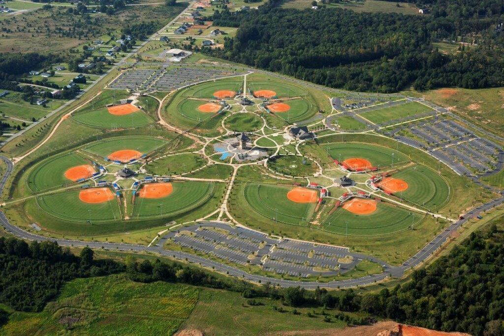 Vista aérea de um complexo esportivo com vários campos de beisebol, áreas de estacionamento e um edifício central.