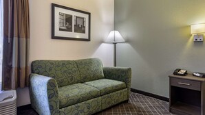 Quarto de hotel com um sofá de cor verde com padrão, um quadro na parede, um candeeiro de pé, uma mesa de cabeceira com telefone e um radiador.