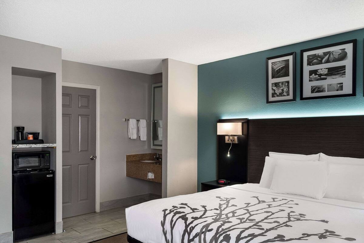 A hotel room with a bed, a nightstand, a wall-mounted lamp, a microwave, a sink, and a door leading to another room.