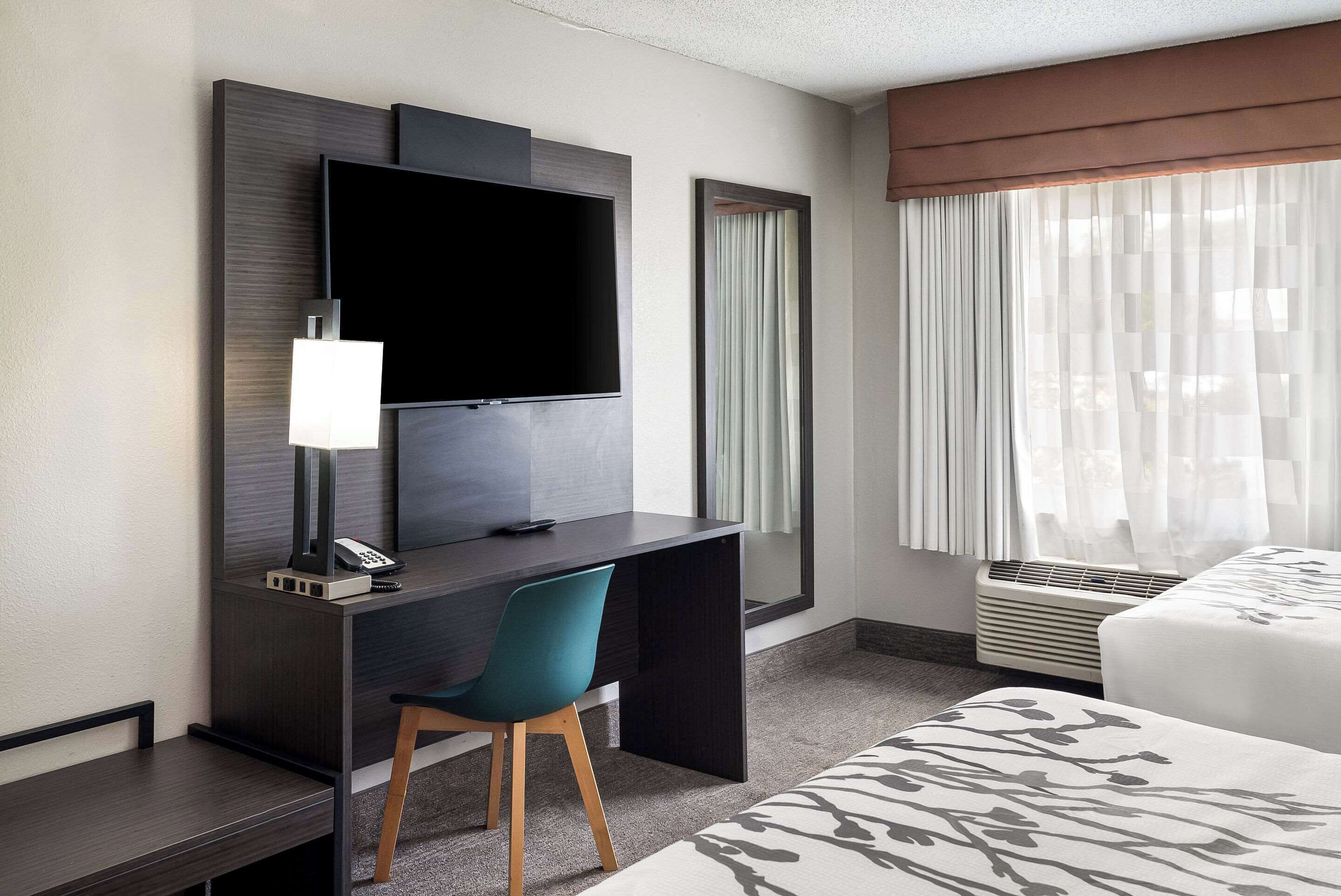 Ein Hotelzimmer mit dunklem Holzschreibtisch, einem Flachbildfernseher, einem türkisfarbenen Sessel, einem Bett mit gemustertem Überwurf und einem großen Fenster mit transparenten Vorhängen.