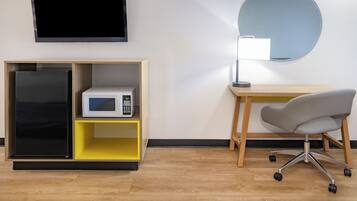 A room with a flat-screen TV mounted on the wall, a microwave on a yellow shelf, a desk with a chair, and a round mirror.