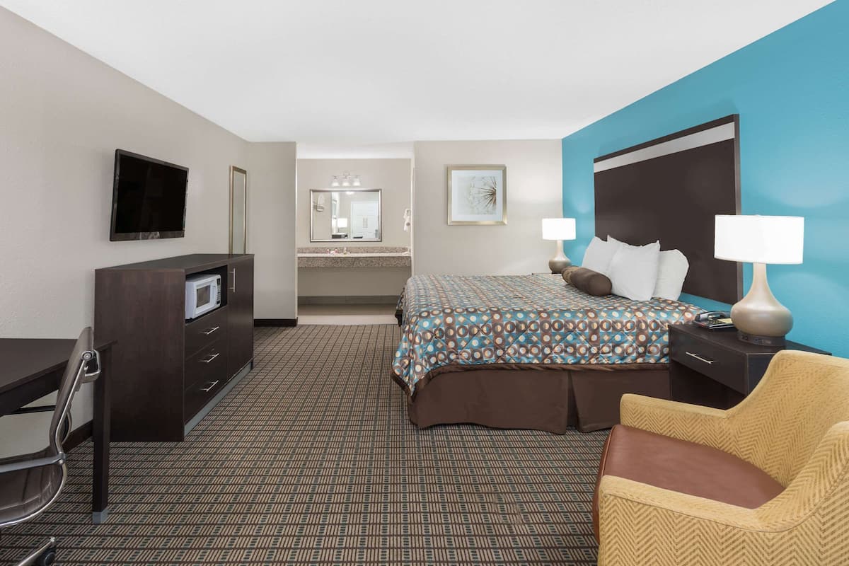 A hotel room with a bed, a desk, a chair, a TV, a mirror, and a bathroom visible in the background.