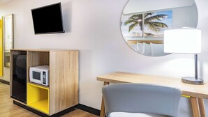 A hotel room with a wooden desk, a grey chair, a microwave, a TV, and a round mirror with a beach painting.