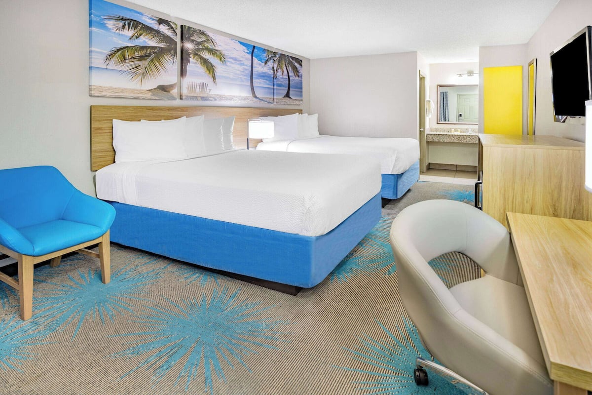 A hotel room with two beds, a blue chair, a bathtub, and a painting of a beach scene.