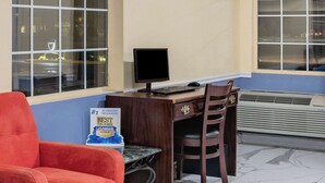 A hotel room with a wooden desk, a computer, a chair, a red armchair, and a sign on the wall.