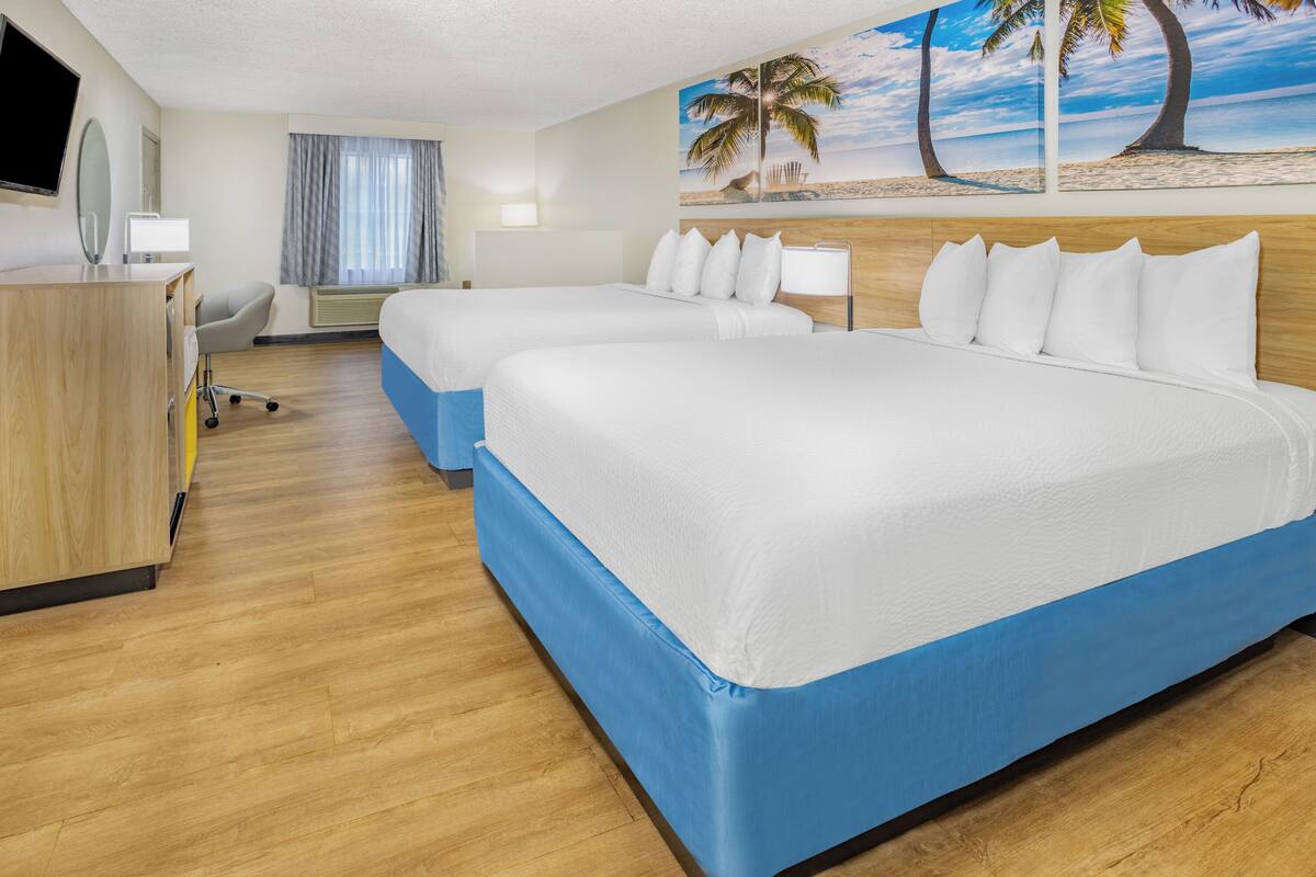 A hotel room with two beds, a desk, a chair, a TV, and a painting of a beach scene.