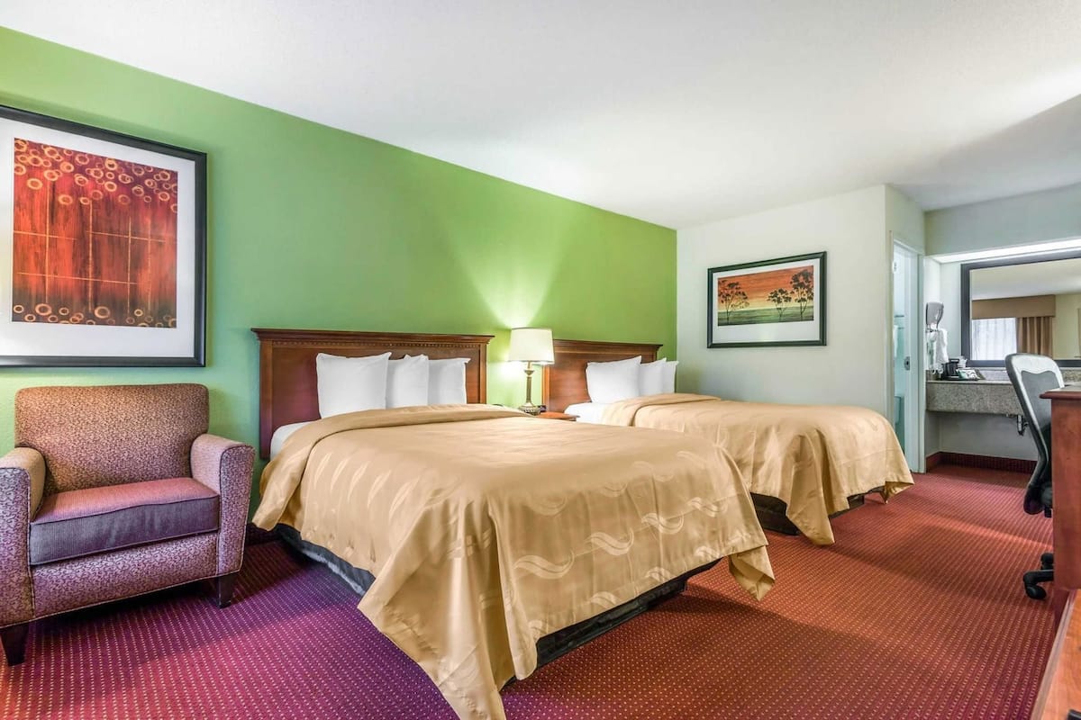 A hotel room with two beds, a chair, a desk, and a bathroom visible through an open door.