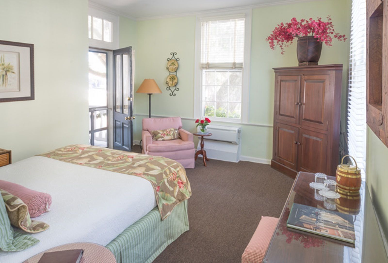 A bedroom with a bed, a chair, a table with a lamp, a cabinet, and a window with flowers.