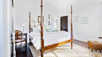 A four-poster bed with a white bedspread, a wooden chair, a wicker chair, and a desk with a lamp.