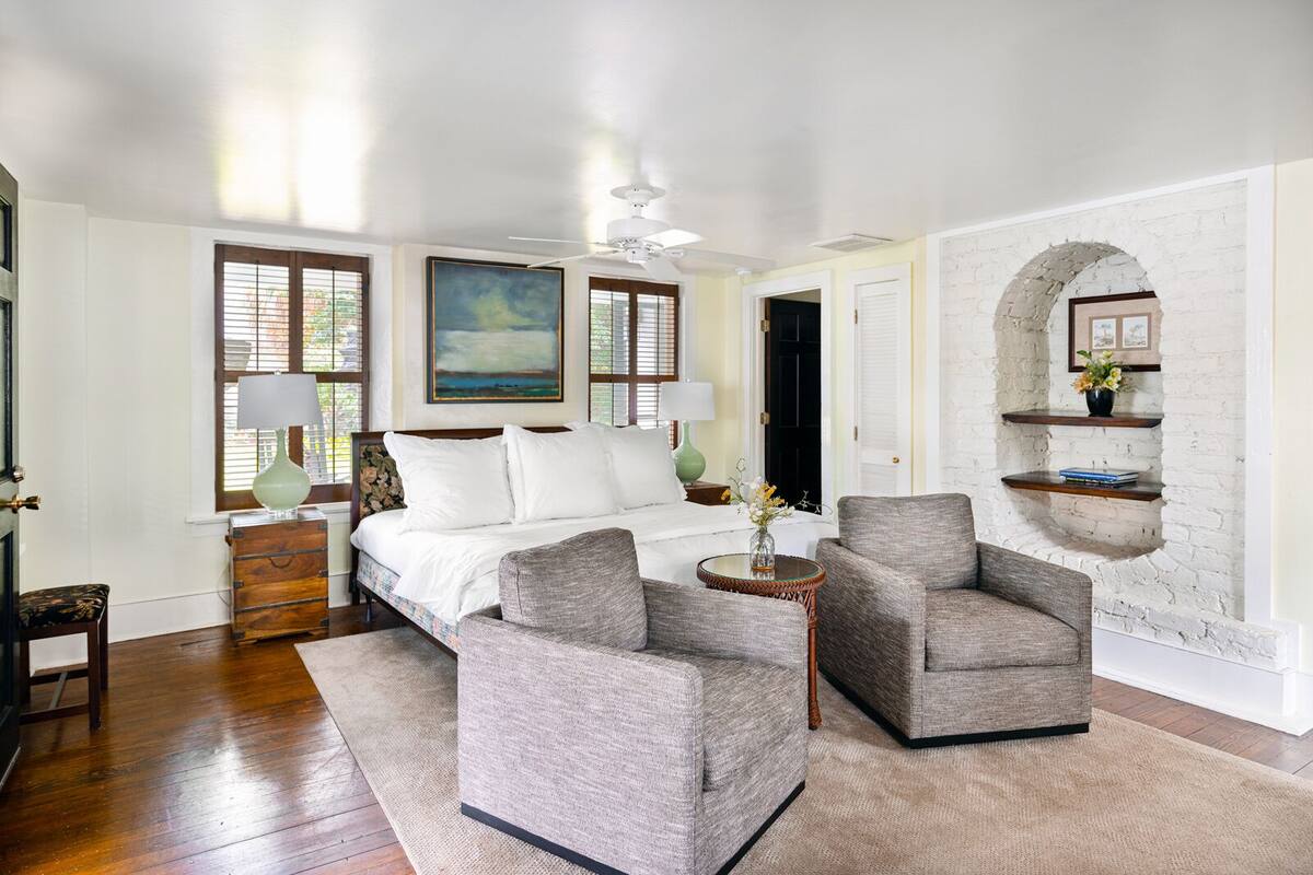 Une chambre à coucher avec un lit, deux fauteuils, une petite table et un mur en briques blanches.
