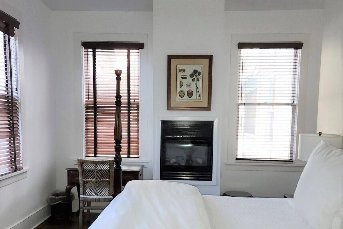 Une chambre à coucher comprenant un lit, une chaise, une cheminée et deux fenêtres équipées de stores.