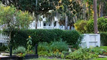 A white house with a porch, surrounded by a garden with various plants and flowers.