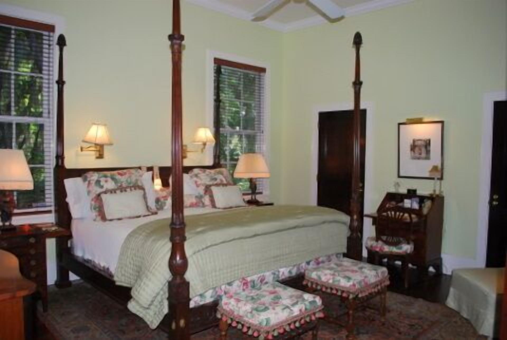 A four-poster bed with floral bedding, two bedside tables with lamps, a wooden wardrobe, and a small round table with chairs.