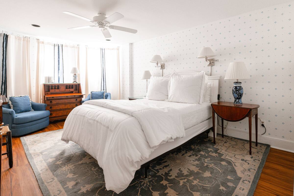 Une chambre à coucher avec un grand lit, un ventilateur de plafond, une commode en bois, un fauteuil bleu et un tapis à motifs.
