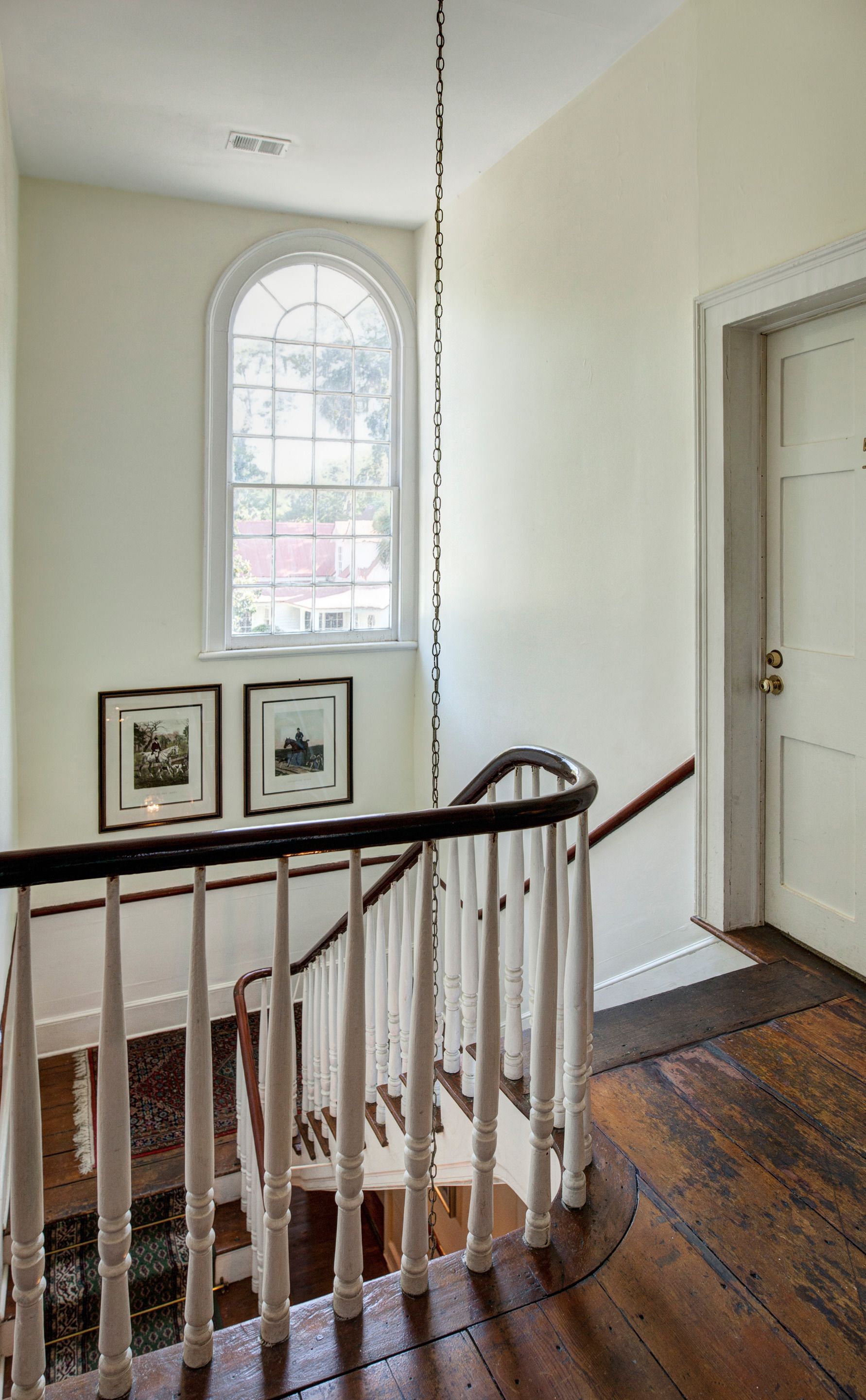 A staircase with white balusters and a wooden handrail, leading to a room with a large arched window and two framed pictures on the wall.