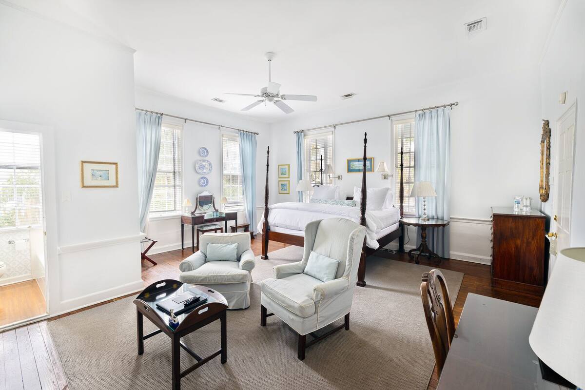 Une chambre spacieuse avec un lit à baldaquin, un coin salon comprenant un fauteuil et une table, ainsi qu’une grande fenêtre avec des rideaux.