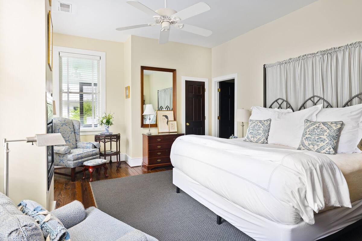 Une chambre à coucher avec un grand lit, une chaise, une commode, un miroir et un ventilateur de plafond.